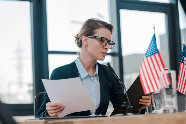 Selektiver Fokus eines attraktiven Diplomaten in Brille, der auf das Klemmbrett in der Nähe amerikanischer Flaggen blickt — Stockfoto