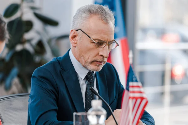 Selektiver Fokus des bärtigen Diplomaten mit Brille in der Nähe amerikanischer Flaggen — Stockfoto