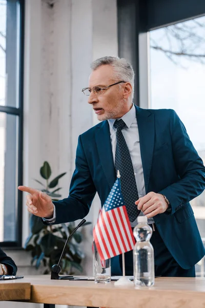 Selective focus of bearded diplomat in eyeglasses pointing with finger while talking near american flag — Stock Photo