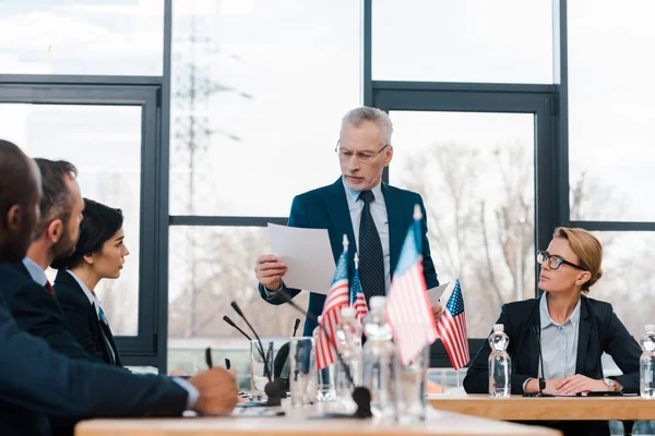Selektiver Fokus des bärtigen Geschäftsmannes, der Papier in der Nähe multikultureller Diplomaten und amerikanischer Flaggen hält — Stockfoto