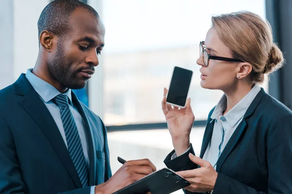 Attractive businesswoman looking at african american businessman and holding smartphone with blank screen — Stock Photo