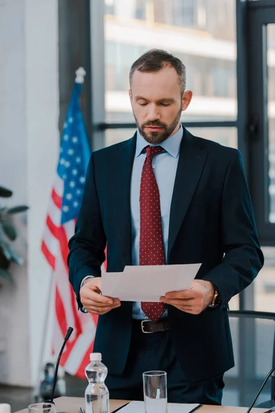 Selective focus of bearded diplomat in suit holding papers near american flag — Stock Photo