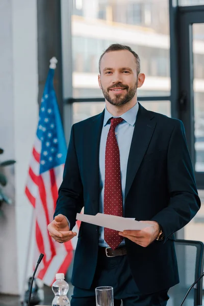 Selective focus of happy diplomat in suit holding papers near american flag — Stock Photo