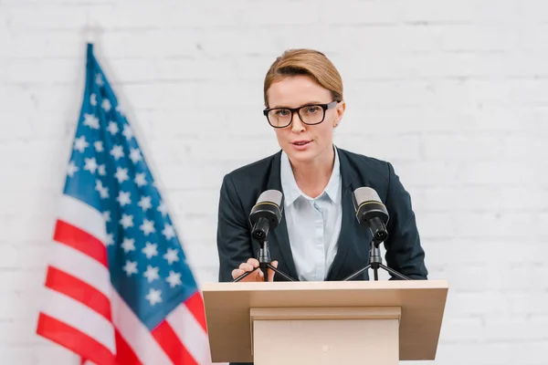 Alto-falante atraente em óculos falando perto de microfones e bandeira americana — Fotografia de Stock
