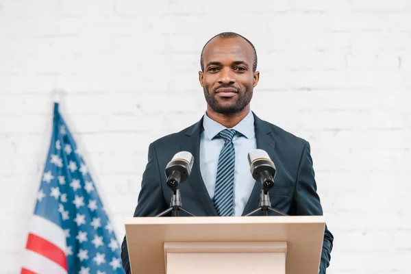 Altavoz afroamericano feliz de pie cerca de micrófonos y bandera americana - foto de stock