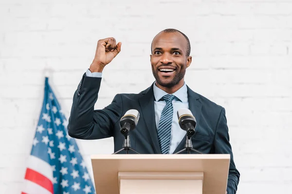 Altavoz afroamericano feliz con puño cerrado sonriendo cerca de micrófonos y bandera americana - foto de stock