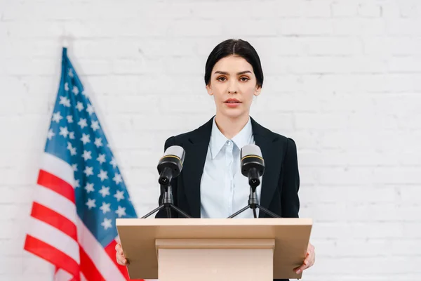 Schöner Redner, der in der Nähe von Mikrofonen und amerikanischer Flagge spricht — Stockfoto