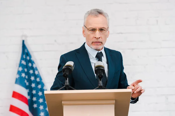 Bärtiger Redner mit Brille, der mit dem Finger in die Nähe von Mikrofonen und amerikanischer Flagge zeigt — Stockfoto