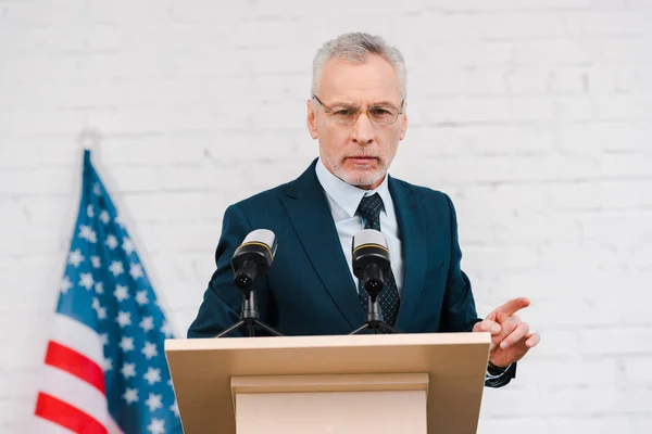 Bärtiger Redner mit Brille, der mit dem Finger in die Nähe von Mikrofonen und amerikanischer Flagge zeigt — Stockfoto