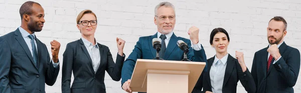 Panoramic shot of happy speaker in glasses and multicultural businessmen and businesswomen showing clenched fists — Stock Photo