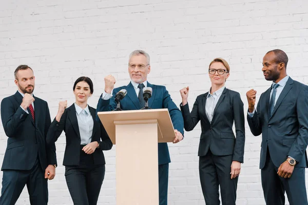 Orador feliz en gafas y hombres de negocios multiculturales y mujeres de negocios mostrando los puños cerrados - foto de stock