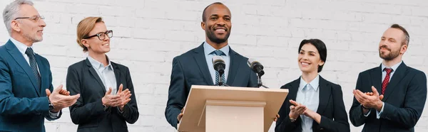 Panoramic shot of businessmen and businesswomen applauding to african american speaker — Stock Photo