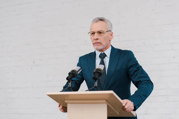 Bearded speaker in glasses standing near microphones and brick wall — Stock Photo