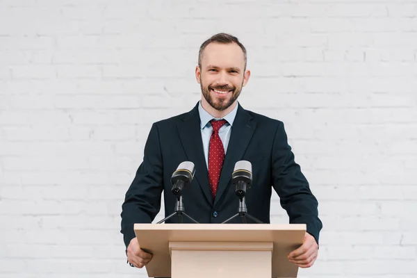 Heureux haut-parleur barbu souriant près des microphones et mur de briques — Photo de stock