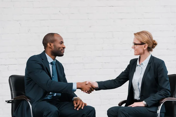 Vista lateral del hombre de negocios afroamericano feliz estrechando la mano con la mujer de negocios mientras está sentado en la silla - foto de stock