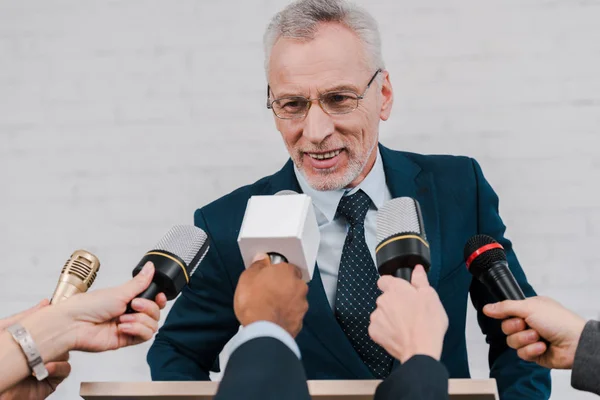 Cropped view of journalists holding microphones near happy bearded diplomat in glasses — Stock Photo