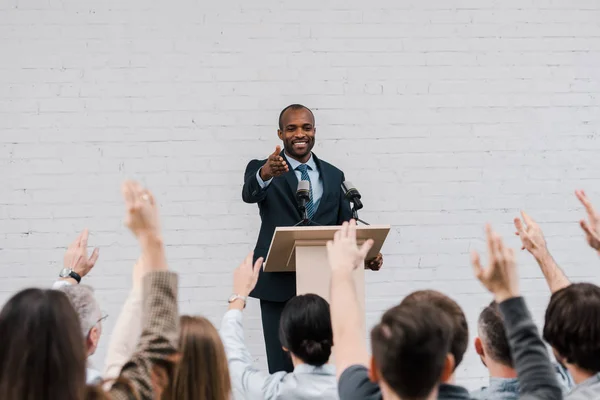 Rückansicht von Journalisten mit erhobenen Händen in der Nähe eines fröhlichen afrikanisch-amerikanischen Sprechers, der neben Mikrofonen gestikuliert — Stockfoto