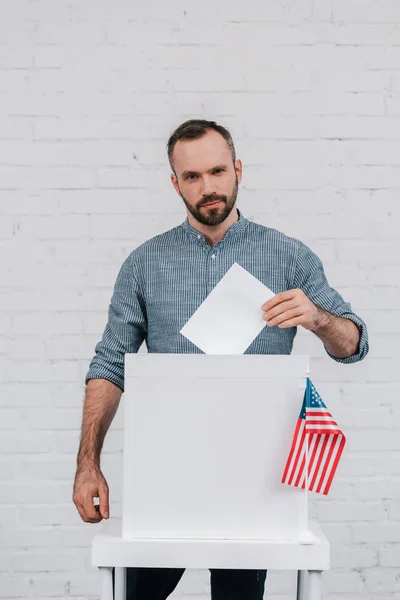 Bärtige und gutaussehende Wähler werfen leere Stimmzettel in die Wahlurne nahe der amerikanischen Flagge — Stockfoto