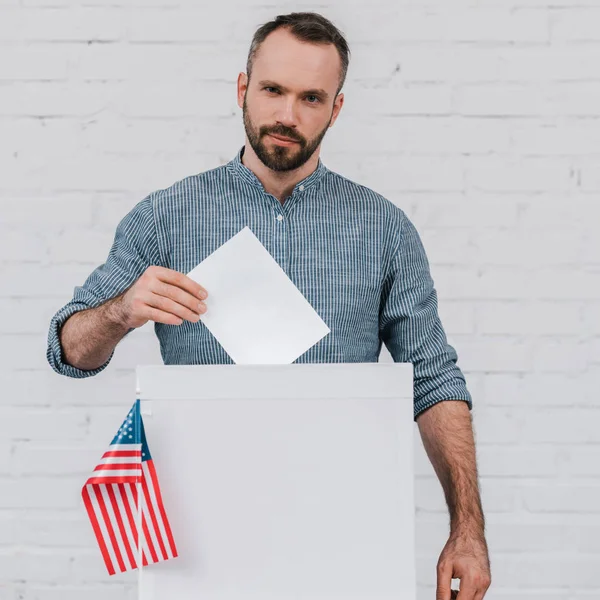 Vista cortada do eleitor colocando cédula em branco na caixa de votação perto da bandeira americana — Fotografia de Stock