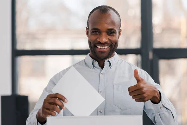 Heureux électeur afro-américain tenant un bulletin blanc et montrant pouce vers le haut — Photo de stock