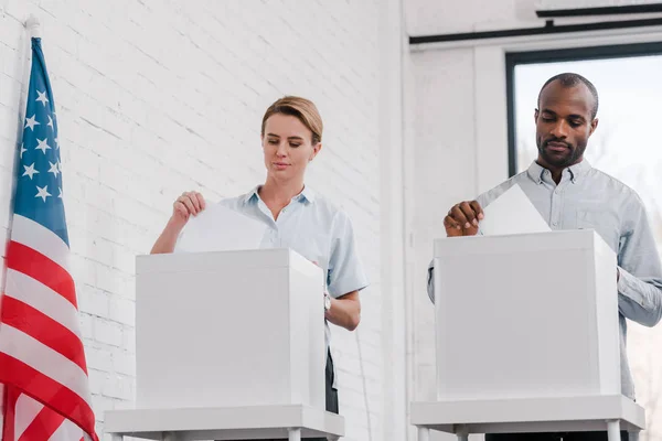 Multikulturelle Bürger werfen Stimmzettel in Wahlurnen nahe der Flagge der USA — Stockfoto