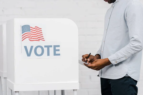 Cropped view of african american citizen holding pen and ballot while voting near stand with vote lettering — Stock Photo