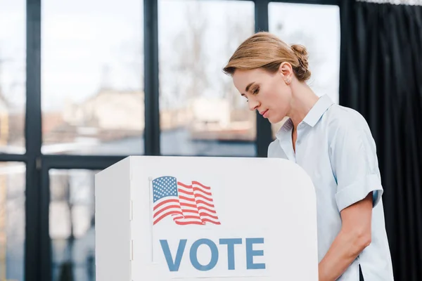 Vue de côté de femme attrayante votant près de stand avec lettrage de vote et drapeau américain — Photo de stock