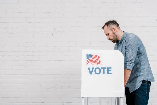 Side view of bearded citizen voting near stand with vote lettering and flag of america — Stock Photo