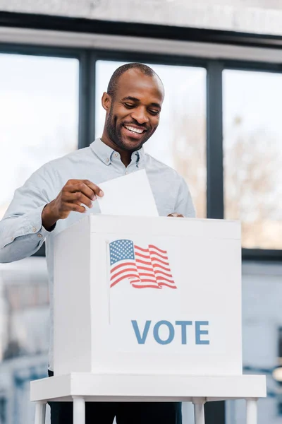 Fröhlicher afrikanisch-amerikanischer Mann bei der Stimmabgabe und beim Einwerfen von Stimmzetteln in eine Urne mit der Flagge Amerikas — Stockfoto