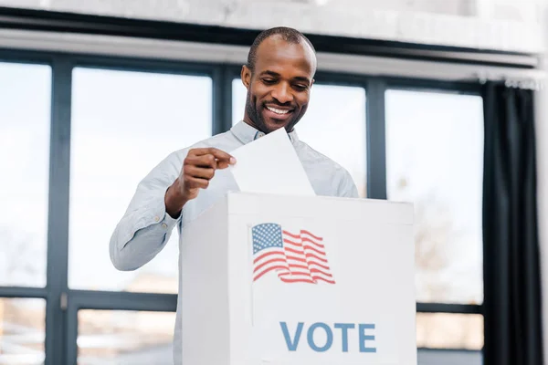 Счастливый африканский американец голосует и кладет бюллетень в коробку с флагом Америки — стоковое фото