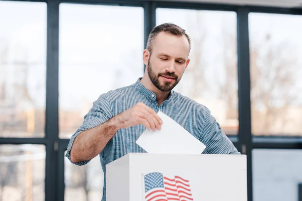 Hombre barbudo votando y poniendo boleta en caja bandera americana - foto de stock