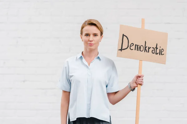 Mulher atraente segurando cartaz com demokratie lettering perto da parede de tijolo — Fotografia de Stock