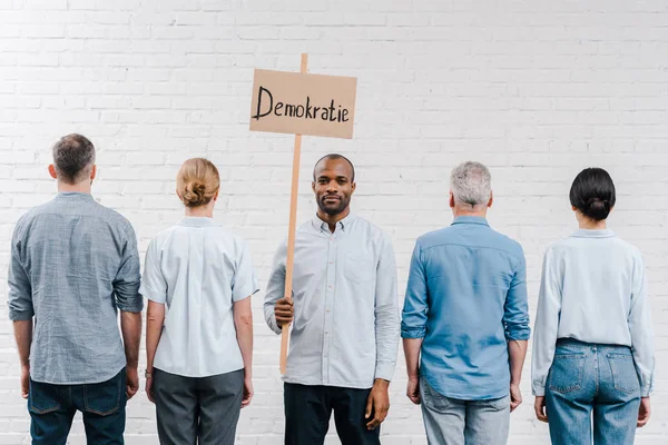 Vista trasera de personas de pie cerca de la pared de ladrillo cerca de hombre afroamericano sosteniendo pancarta con letras demokratie - foto de stock