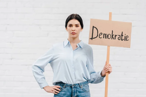 Mulher segurando cartaz com demokratie letras e de pé com a mão no quadril perto da parede de tijolo — Fotografia de Stock