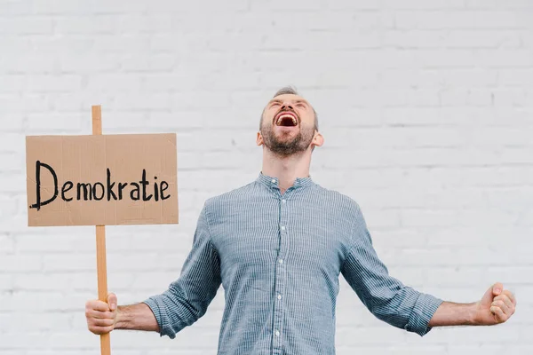 Hombre emocional sosteniendo pancarta con letras demokratie y gritando cerca de la pared de ladrillo - foto de stock