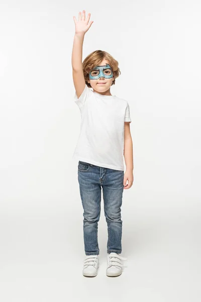 Vista completa de niño sonriente con máscara de superhéroe pintado en la cara de pie con la mano levantada sobre fondo blanco - foto de stock