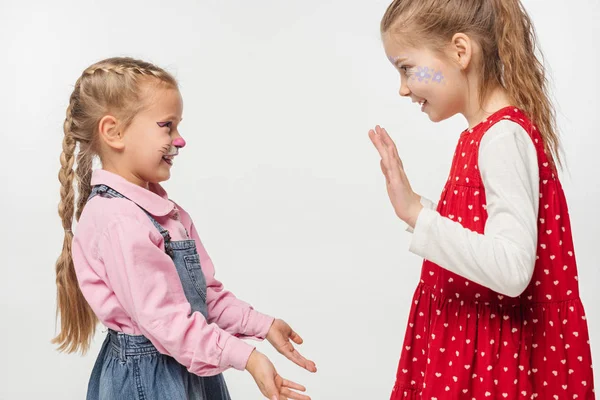 Amigos emocionados con bozal de gato y pinturas florales en caras aplaudiendo las manos entre sí aislados en blanco - foto de stock