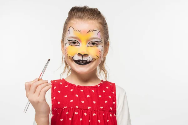 Smiling child with tiger muzzle painting on face looking at camera while holding paintbrush isolated on white — Stock Photo