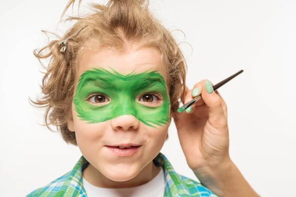 Cropped view of artist painting gecko mask on face of shaggy, cheerful boy isolated on white — Stock Photo