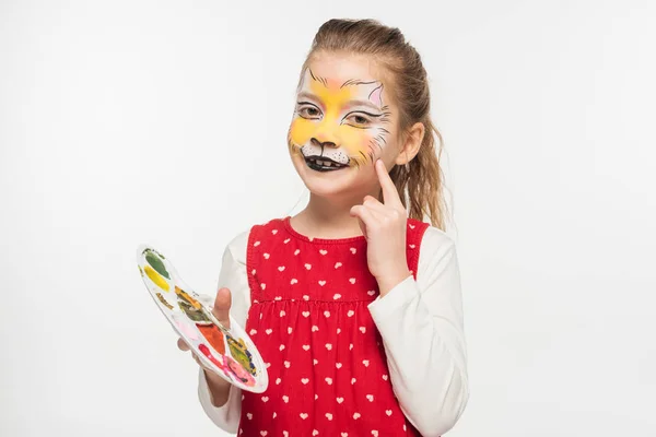 Bambino sorridente con la pittura muso di tigre sulla tavolozza di tenuta del viso e puntando con il dito al viso isolato su bianco — Foto stock