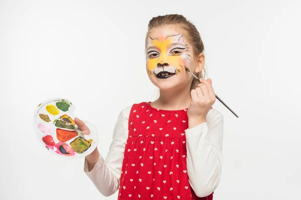 Lindo niño con tigre bozal pintura en la cara celebración de la paleta mientras dibuja en la mejilla con pincel aislado en blanco - foto de stock