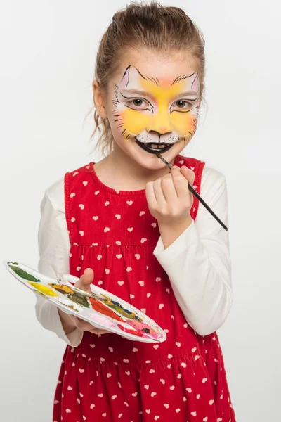 Garoto sorridente com pintura de focinho de tigre na paleta de rosto enquanto pinta nos lábios com pincel isolado no branco — Fotografia de Stock