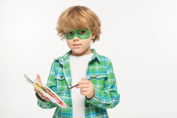 Adorable niño con máscara de geco pintado en la cara celebración de la paleta y el pincel mientras mira la cámara aislada en blanco - foto de stock