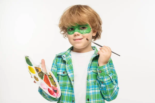 Niño alegre con máscara de geco pintado en la cara tocando mejilla con pincel aislado en blanco - foto de stock