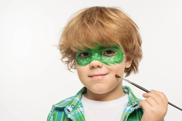 Carino ragazzo con maschera di geco dipinta sul viso toccare guancia con pennello isolato su bianco — Foto stock