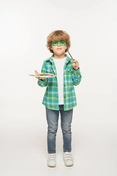 Full length view of cute boy with gecko mask painted on face holding palette and paintbrush on white background — Stock Photo