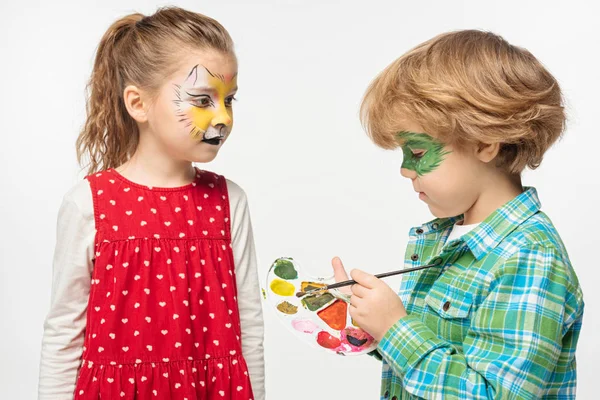 Adorable niño con máscara de geco pintado celebración paleta y pincel cerca amigo con tigre bozal pintura en la cara aislada en blanco - foto de stock