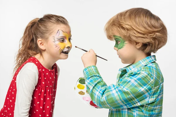 Lindo chico con máscara de geco pintado celebración paleta y pincel cerca de amigo con tigre bozal pintura en la cara aislada en blanco - foto de stock