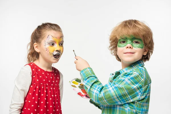 Joyeux garçon avec masque de gecko peint tenant palette et pinceau ami proche avec tigre museau peinture sur le visage isolé sur blanc — Photo de stock