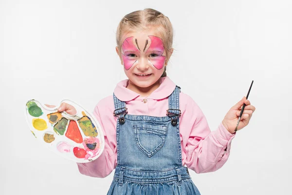 Lächelndes Kind mit Schmetterlingsmalerei im Gesicht, Palette und Pinsel in der Hand, während es die Kamera isoliert auf Weiß betrachtet — Stockfoto